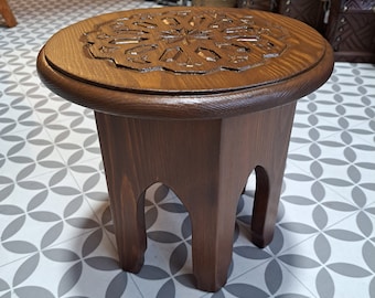 May include: A small, round wooden stool with a carved design on the top. The stool has three legs and is made of dark brown wood.