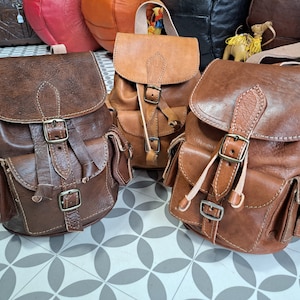 May include: Three brown leather backpacks with adjustable straps and buckles. Each backpack has two front pockets.