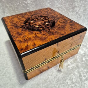 May include: A wooden box with a burl wood top and sides. The top features a decorative carved design and is framed with black trim. The box has a gold-coloured tassel attached to the keyhole.