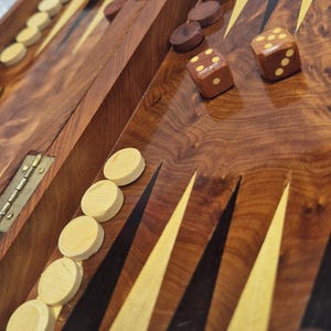 May include: A wooden backgammon set with a rich, dark wood grain finish. The game board features alternating black, yellow, and brown points. Dice and wooden game pieces are scattered across the board, ready for play.