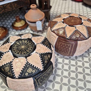 May include: Two Moroccan poufs, one black and white with gold accents and one brown and white with gold accents. The poufs are made of leather and have a traditional Moroccan design.