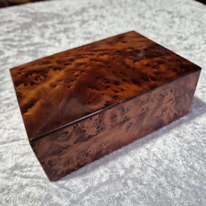 May include: A wooden box with a rich brown burl wood finish. The box has a hinged lid and is made of solid wood.