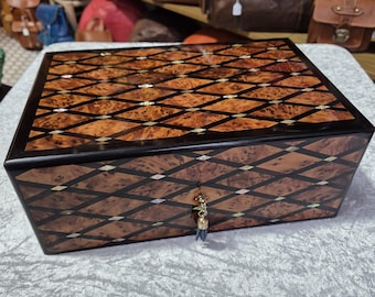 Puede incluir: Una caja de madera con un diseño de incrustaciones de diamantes decorativos. La caja está hecha de madera oscura con incrustaciones de color marrón y dorado. La caja tiene un pequeño borla que cuelga de la cerradura.