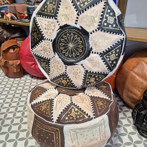 May include: Two round, leather poufs with intricate geometric patterns in black, white, and gold. The poufs are stacked on top of each other.