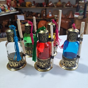 May include: Five small glass bottles with decorative metal tops and colourful liquids inside. Each bottle has a wooden stick and a tassel hanging from the top. The bottles are sitting on a white surface.