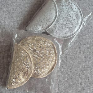 Op de afbeelding: Twee paar bh-inzetstukken in een doorzichtige plastic verpakking. Eén paar is zilverkleurig, de andere goudkleurig. Beide hebben een textuurpatroon en gestikte randen. De inzetstukken zijn voor in de bh.