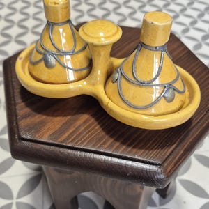 Puede incluir: Un juego de condimentos de cerámica amarilla con un asa central, con dos recipientes con tapa y un pequeño cuenco central. El juego está decorado con detalles metálicos plateados y se asienta sobre un soporte hexagonal de madera oscura.