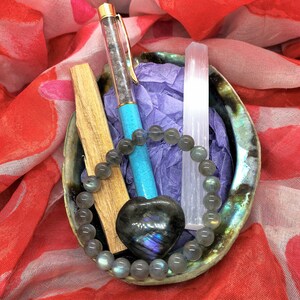 Peut inclure: Un bol en coquille de mer rempli de cristaux et un stylo turquoise. Le bol est posé sur un tissu à motifs rouge et blanc. Les cristaux comprennent un cœur de labradorite, une baguette de cristal clair et un bracelet de perles grises.
