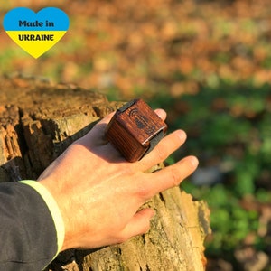 Può includere: Un anello di legno con un elastico nero e una piccola scatola di legno attaccata. La scatola ha un disegno sulla parte superiore e le parole "Made in Ukraine" sono stampate su un adesivo a forma di cuore. L'anello è sul dito di una persona.