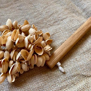 Puede incluir: Una herramienta de cocina artesanal. Presenta un grupo de cáscaras de pistacho de color marrón claro unidas a un mango de madera clara. El mango tiene una pequeña cuerda blanca atada al final. Fondo texturizado.