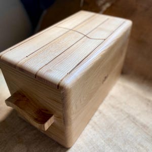 May include: A light-colored wooden box with a handle on the side. The top of the box has carved lines. The wood grain is visible. The box is rectangular with rounded edges.