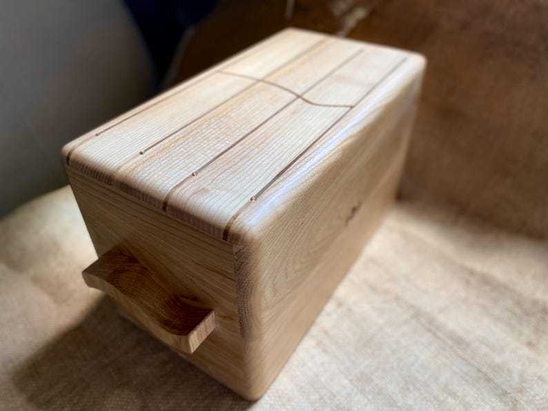 May include: A light-colored wooden box with a handle on the side. The top of the box has carved lines. The wood grain is visible. The box is rectangular with rounded edges.