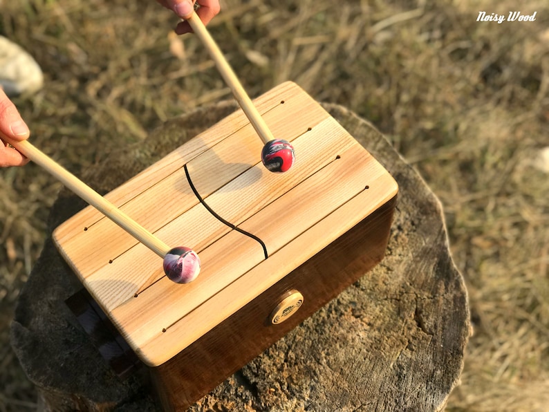 May include: A wooden tongue drum with two mallets. The drum has a light wood top with carved sound slots and a dark brown base. The mallets have wooden handles and colorful, patterned heads. The text "Noisy Wood" is in the upper right corner.