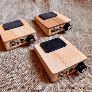 May include: Three rectangular wooden stompboxes with light wood grain, each featuring a black rectangular top pad, a silver input jack, a black control knob, and a small logo printed on the side panel. The devices are arranged on a textured surface.