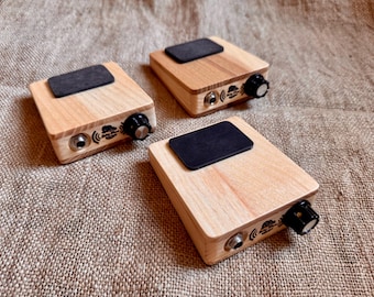 May include: Three rectangular wooden stompboxes with light wood grain, each featuring a black rectangular top pad, a silver input jack, a black control knob, and a small logo printed on the side panel. The devices are arranged on a textured surface.