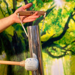 Puede incluir: Un carillón de viento cromado con un percutor de madera y un mango de madera. El carillón es cilíndrico y tiene un pequeño agujero. El percutor tiene una cabeza azul y blanca. El mango tiene el texto "Singing Tree".