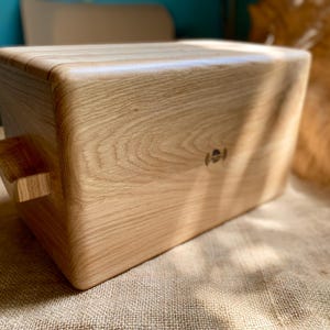 May include: A rectangular wooden box with rounded corners and a handle on one side. The box is made of light-colored wood, showcasing the natural grain. A small, circular emblem is visible on the side. The box sits on a textured, tan surface.
