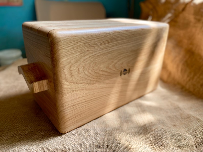 May include: A rectangular wooden box with rounded corners and a handle on one side. The box is made of light-colored wood, showcasing the natural grain. A small, circular emblem is visible on the side. The box sits on a textured, tan surface.
