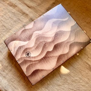 May include: A rectangular wooden cajon drum featuring a smooth, wavy, multi-tonal brown and cream gradient pattern across its front face. The surface is secured with small silver screws along the perimeter and displays a circular Noisy Wood logo.