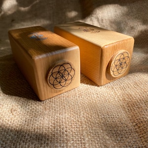 May include: Two rectangular wooden blocks with rounded edges. Each block has a circular design on one end, featuring a black geometric pattern. The wood has a light, natural colour, and the blocks are resting on a textured, tan surface.