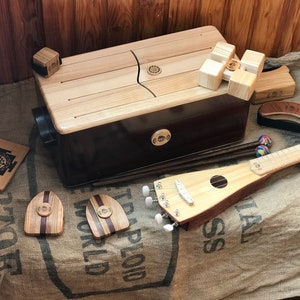 May include: A collection of wooden musical instruments, including a large rectangular kalimba, a small ukulele-like instrument, and various percussion pieces. The instruments are made of light and dark wood, with a natural finish. The instruments are displayed on a burlap surface.