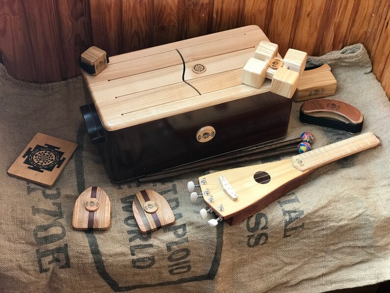 May include: A collection of wooden musical instruments, including a large rectangular kalimba, a small ukulele-like instrument, and various percussion pieces. The instruments are made of light and dark wood, with a natural finish. The instruments are displayed on a burlap surface.