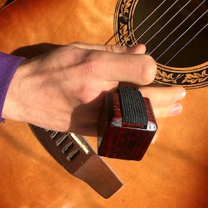 Puede incluir: Primer plano de un dedo con una púa de dedo de madera unida a él. La púa de dedo es de color rojo oscuro con elástico negro y se está utilizando en una guitarra.