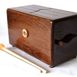 May include: A wooden tongue drum with a rich, dark brown finish. The instrument has a rectangular shape with a handle on one side and a series of slots on the top. Two orange-tipped mallets rest in front of the drum.