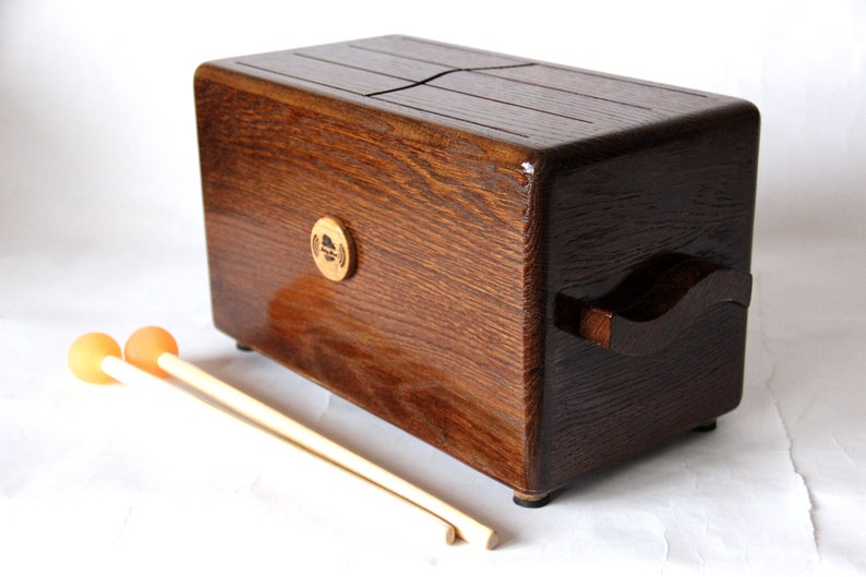 May include: A wooden tongue drum with a rich, dark brown finish. The instrument has a rectangular shape with a handle on one side and a series of slots on the top. Two orange-tipped mallets rest in front of the drum.