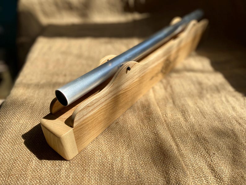 May include: A silver metal tube resting in a wooden stand. The stand is made of light brown wood with a natural finish. The tube is slightly angled and the stand is resting on a brown burlap surface.