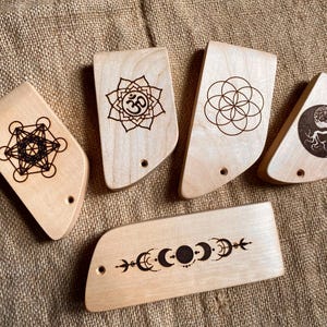 May include: Five wooden items with etched designs, including a Metatron's Cube, an Om symbol, a Flower of Life, a Yin Yang tree, and a crescent moon phase. Each piece has a small hole. The items are made of light-colored wood.