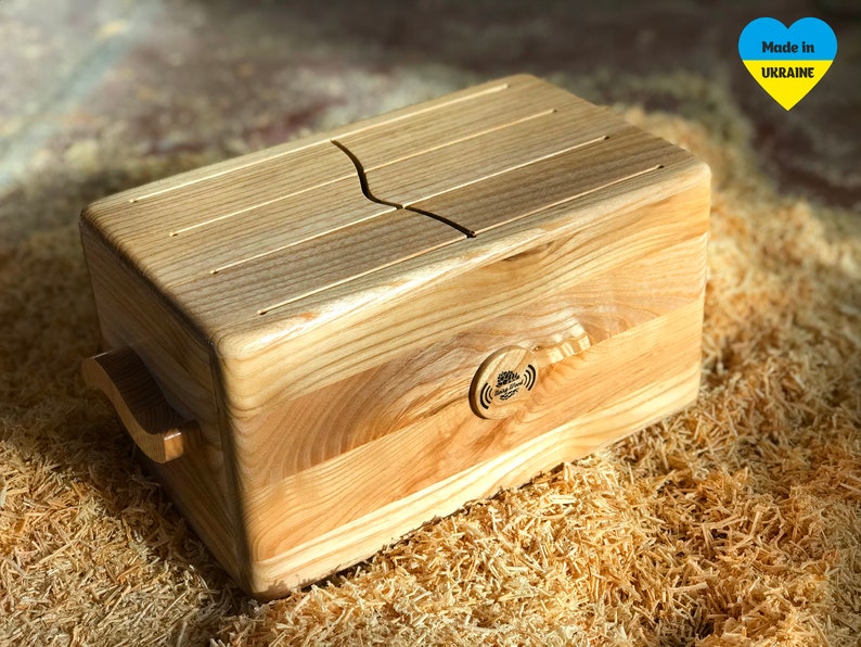May include: A wooden tongue drum with a natural finish. The instrument has a rectangular shape with a curved slot on the top and a handle on the side. A small circular logo is visible on the front. The drum is made in Ukraine.