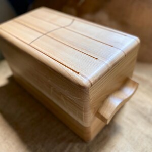 May include: A wooden tongue drum with a natural finish. The instrument has a rectangular body with rounded corners and a curved handle. The top surface features carved tongues for sound production.