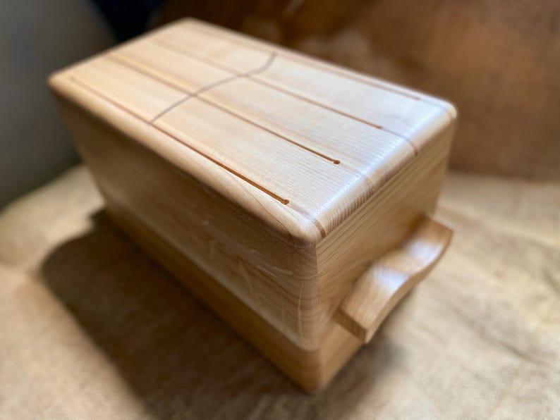 May include: A wooden tongue drum with a natural finish. The instrument has a rectangular body with rounded corners and a curved handle. The top surface features carved tongues for sound production.