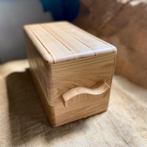 May include: A light-colored wooden bread box with a rectangular shape and a curved handle. The lid has grooves, and the box is made of light-colored wood. The bread box is sitting on a burlap surface.