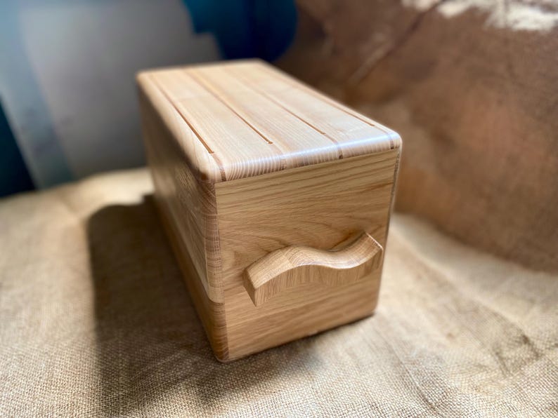 May include: A light-colored wooden bread box with a rectangular shape and a curved handle. The lid has grooves, and the box is made of light-colored wood. The bread box is sitting on a burlap surface.