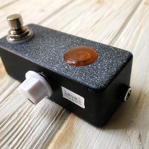 Könnte beinhalten: Ein schwarzes und graues Effektpedal mit einem silbernen Knopf, einem weißen Knopf und einer orangefarbenen Kontrollleuchte. Das Pedal hat eine strukturierte Oberfläche und ein Etikett mit der Aufschrift "Level Noise".