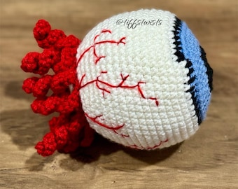 May include: Crocheted eye with a white sclera, blue and black iris, and red veins. The optic nerve is a cluster of red crochet. The eye is a handmade craft item.