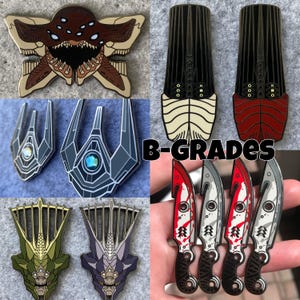 Op de afbeelding: Een verzameling emaille pins in diverse ontwerpen. Inclusief een bruine en tan monster pin, twee zwart-witte pins, twee grijze pins met een blauwe edelsteen en vier mes pins met rode accenten. De tekst "B-GRADES" is ook zichtbaar.