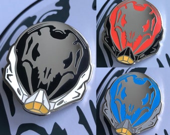 Op de afbeelding: Drie emaillepins met een zwart-wit schedelontwerp met een zilveren afwerking. De pins zijn in drie verschillende kleuren: zwart, rood en blauw.