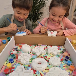 Puede incluir: Niños pintando figuras de yeso con pinceles y pinturas coloridas. El kit incluye varias formas como un coche, una mariposa, una flor y una rosquilla. Las figuras son blancas y las pinturas están en pequeños recipientes coloridos.