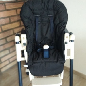 Może przedstawiać: Czarno-białe krzesełko do karmienia z wyściełanym siedziskiem i pasami bezpieczeństwa. Krzesełko do karmienia ma podnóżek i jest podparte czterema nogami. Rama jest biała i granatowa.