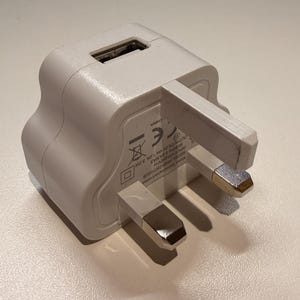 Puede incluir: Un cargador de pared USB blanco con un enchufe de tres clavijas. El cargador tiene un puerto USB y est&aacute; dise&ntilde;ado para dispositivos electr&oacute;nicos. Las clavijas del enchufe tienen puntas plateadas.