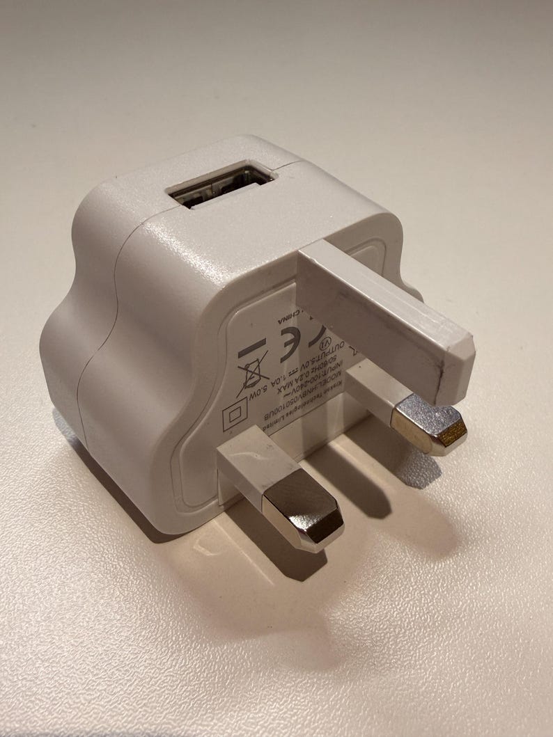 Puede incluir: Un cargador de pared USB blanco con un enchufe de tres clavijas. El cargador tiene un puerto USB y est&aacute; dise&ntilde;ado para dispositivos electr&oacute;nicos. Las clavijas del enchufe tienen puntas plateadas.