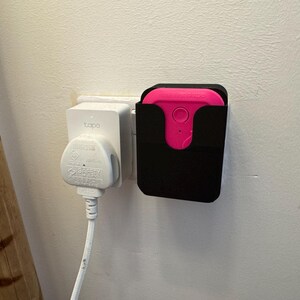 May include: A white electrical outlet with a white plug and cord. Next to it is a black wall mount with a pink device inside.