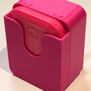 Puede incluir: Una funda rosa brillante, impresa en 3D, con un dispositivo rosa a juego en su interior. El dispositivo tiene un bot&oacute;n circular y el texto "octopus energy" impreso. La funda tiene una tapa con bisagras y una ranura para sujetar el dispositivo.