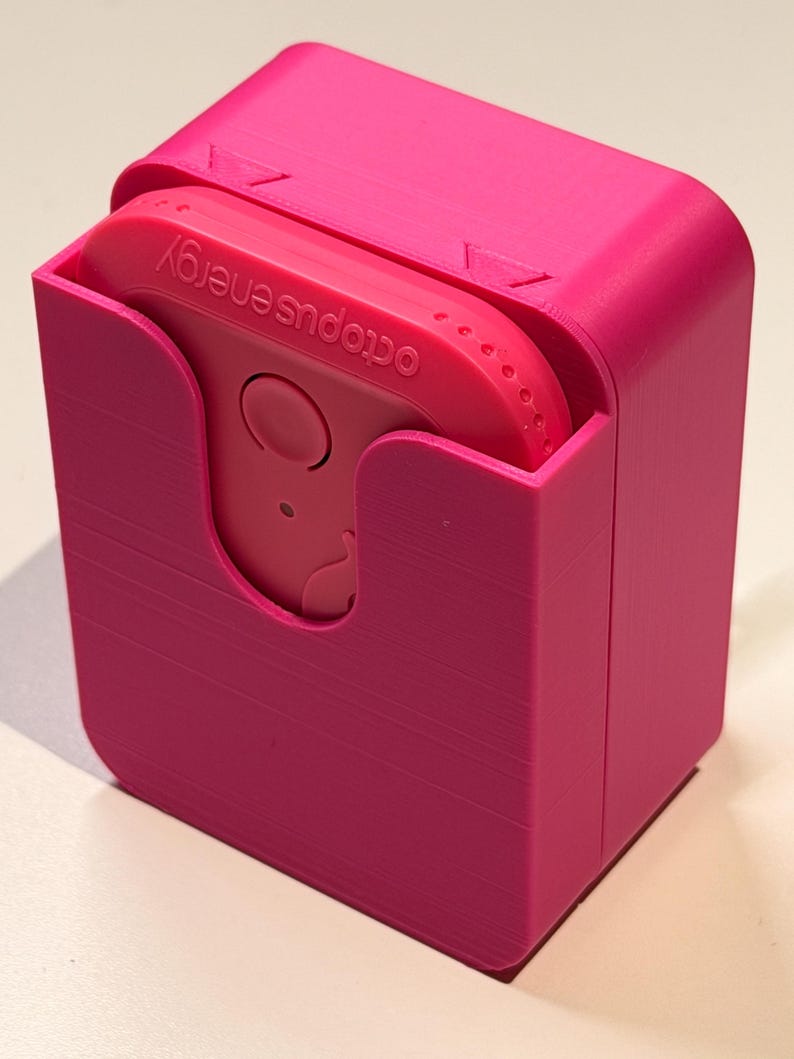Puede incluir: Una funda rosa brillante, impresa en 3D, con un dispositivo rosa a juego en su interior. El dispositivo tiene un bot&oacute;n circular y el texto "octopus energy" impreso. La funda tiene una tapa con bisagras y una ranura para sujetar el dispositivo.