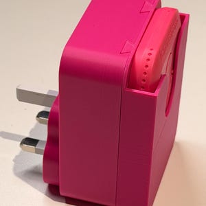 Puede incluir: Un adaptador el&eacute;ctrico rosa brillante con un enchufe de tres clavijas. El adaptador tiene una carcasa rectangular con una ranura para un dispositivo m&aacute;s peque&ntilde;o y redondeado. El dispositivo tambi&eacute;n es rosa y tiene la palabra "Galestron" impresa.