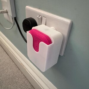 May include: A white plastic wall mount for a pink smart home device. The mount is plugged into a standard electrical outlet.