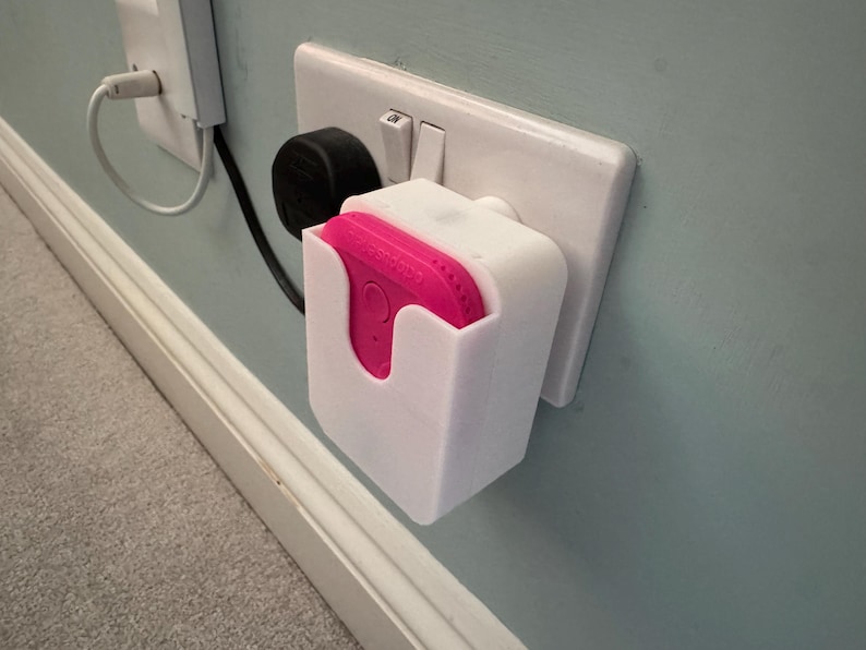 May include: A white plastic wall mount for a pink smart home device. The mount is plugged into a standard electrical outlet.
